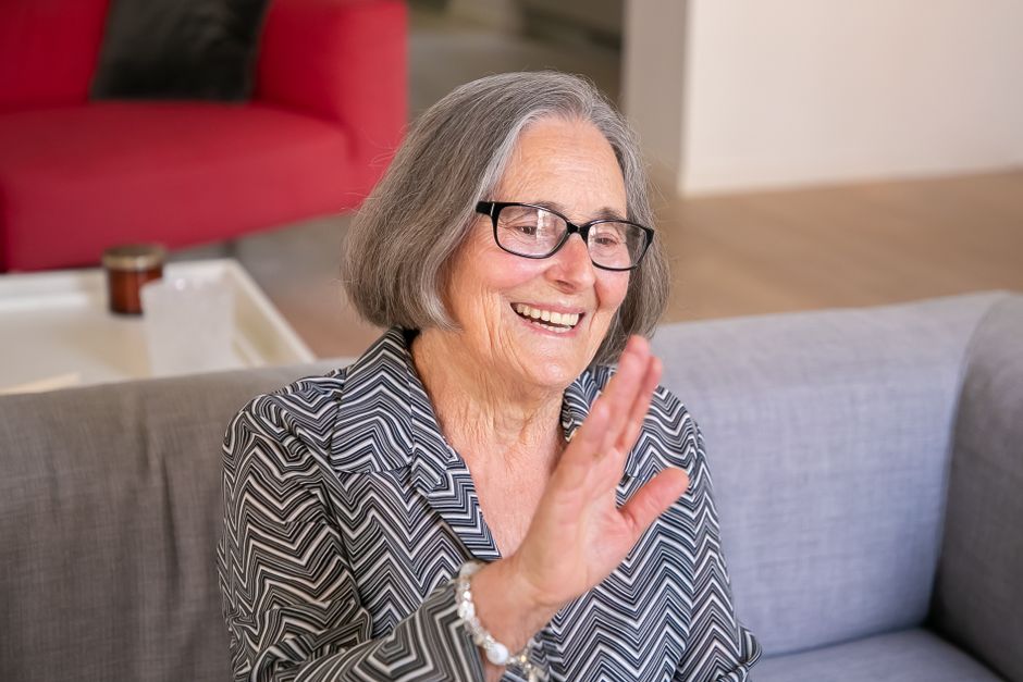 Smiling senior woman with glasses waving while sitting on a couch indoors.
