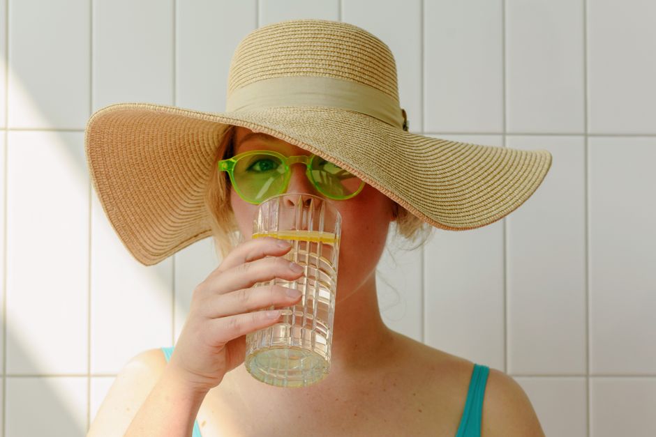 Adult woman in green sunglasses drinking water with lemon slice, wearing a hat.