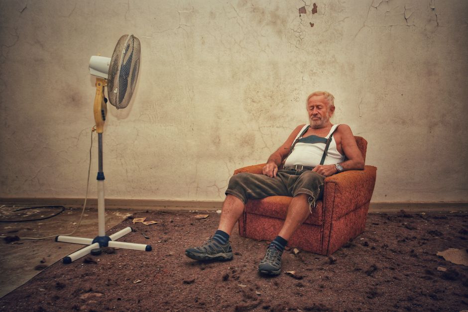 Elderly man resting in an armchair with a fan in a rustic indoor setting.