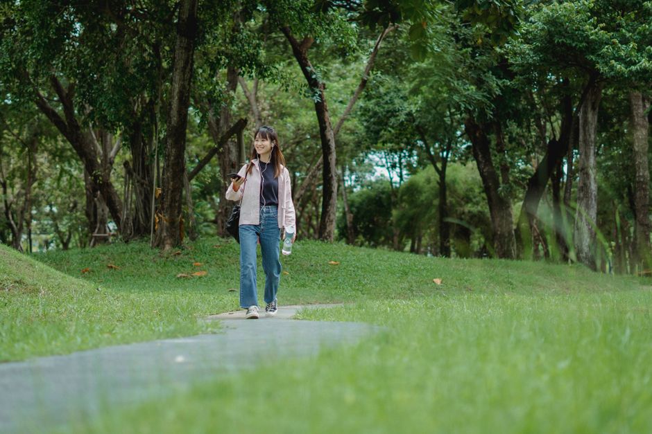 Full length of cheerful young Asian female in casual outfit strolling along green park and talking on phone using earphones