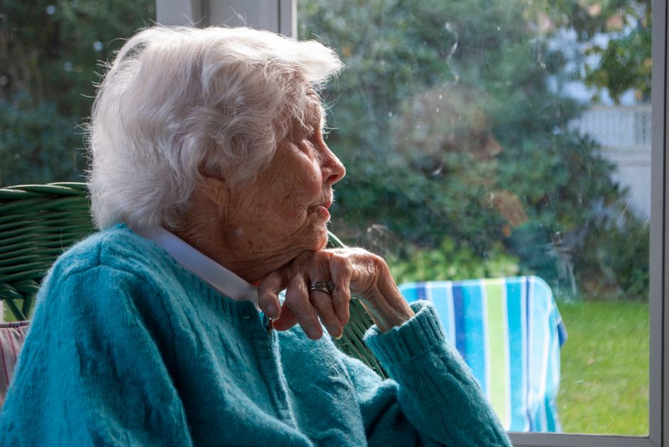 Side view of pensive senior female with white hair in warm sweater leaning on hand and looking away while resting on chair near window at home