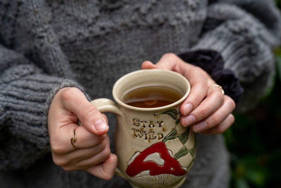A person in a sweater holds a ceramic mug with tea, featuring 'Stay Wild' text, outdoors.