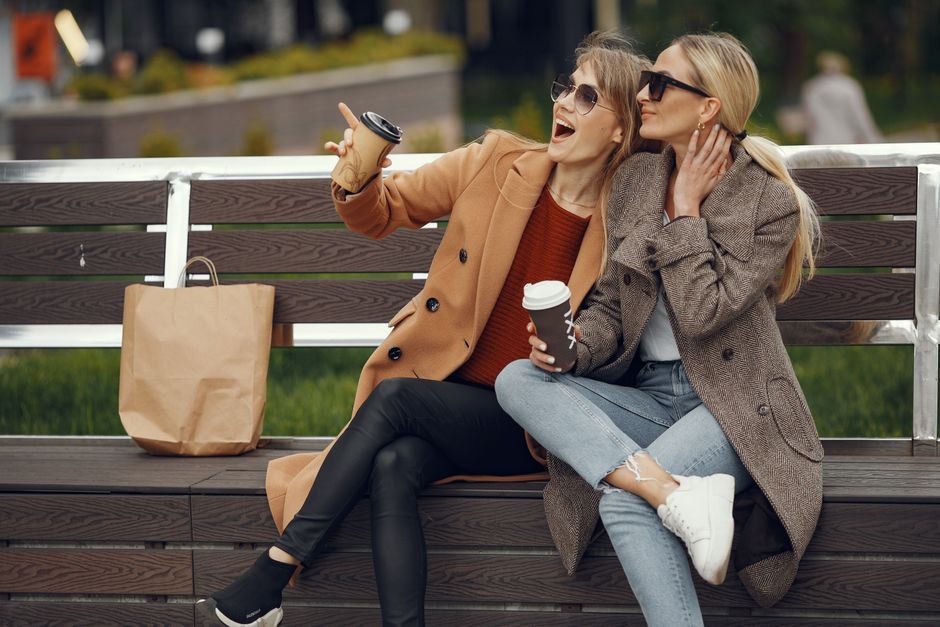 Two women in warm coats share a laugh over coffee on a park bench in autumn.