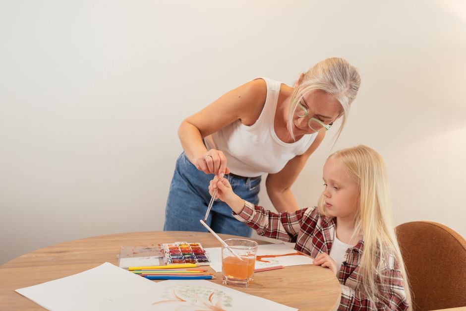 A grandmother guides her granddaughter in watercolor painting at a cozy indoor setting.