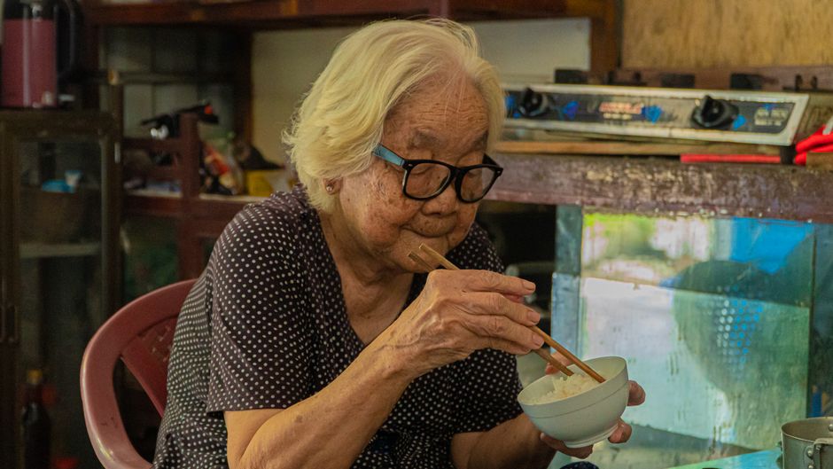Senior Asian woman eating rice with chopsticks in a cozy indoor setting.