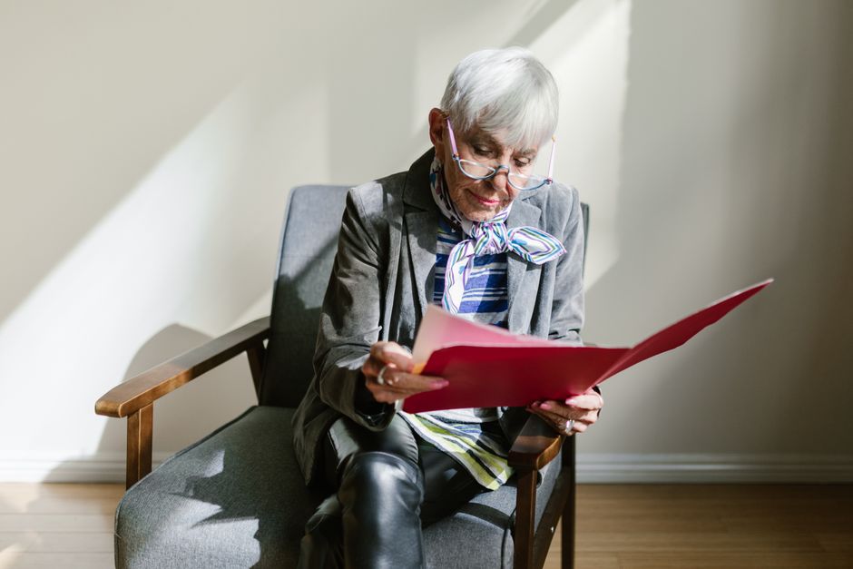 Senior woman in glasses reading a document indoors, seated in an armchair.