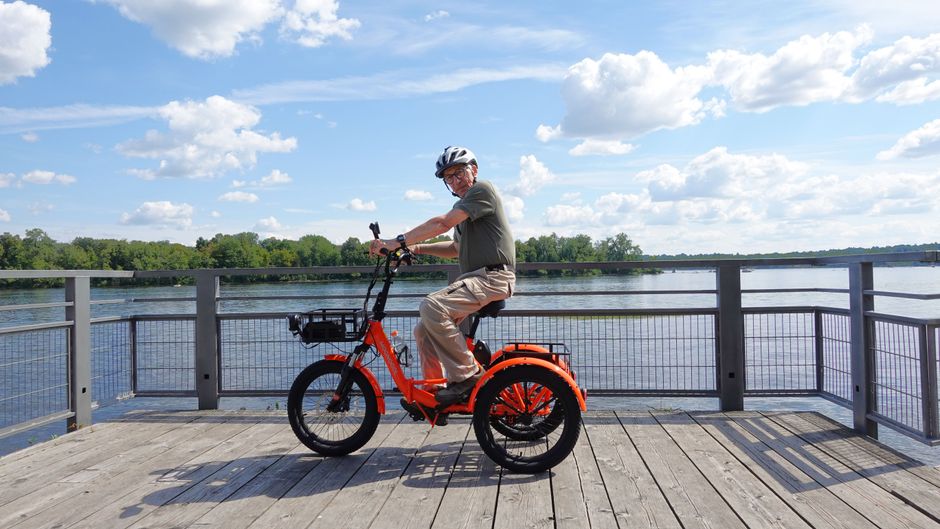Elderly man enjoying a ride on an electric tricycle by the waterfront on a sunny day.