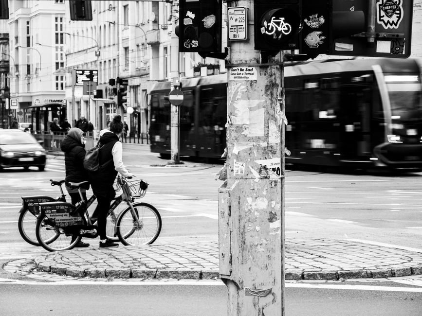 Black and white photo of two cyclists waiting at a busy urban intersection.